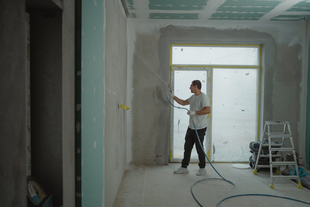 Photo by Tima Miroshnichenko A man sprays paint on a wall while renovating an indoor room with professional equipment.